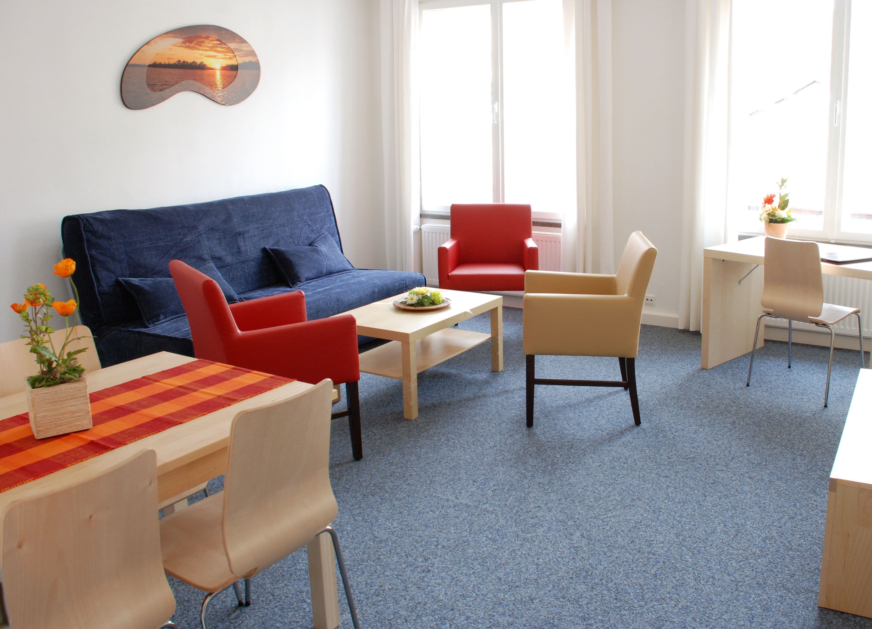 A modern living room with a blue sofa, red chairs, a wooden table, and a small potted plant.