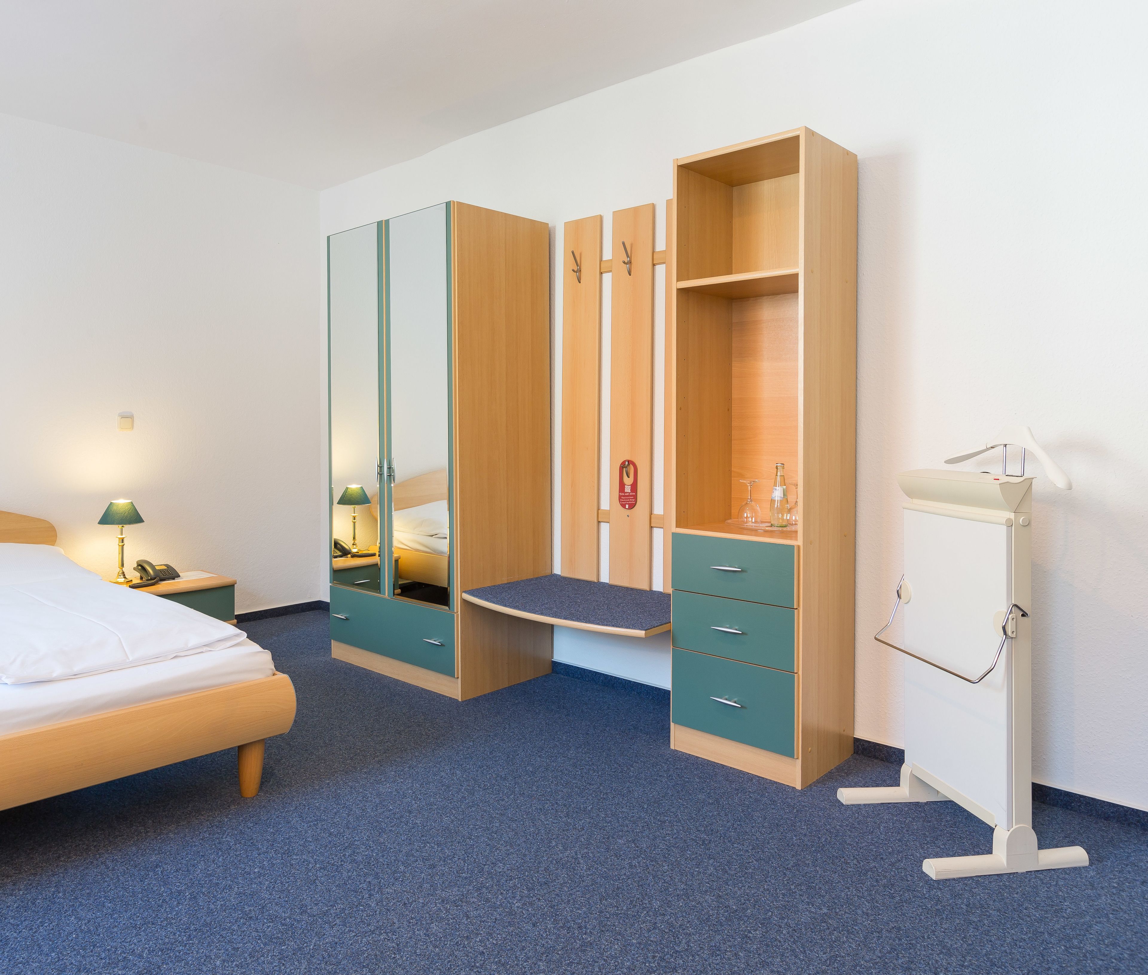Ein Hotelzimmer mit einem Bett, einem Schreibtisch mit Lampe, einem Spiegel, einem Kleiderschrank mit Spiegel und einer kleinen Kommode mit Lampe.