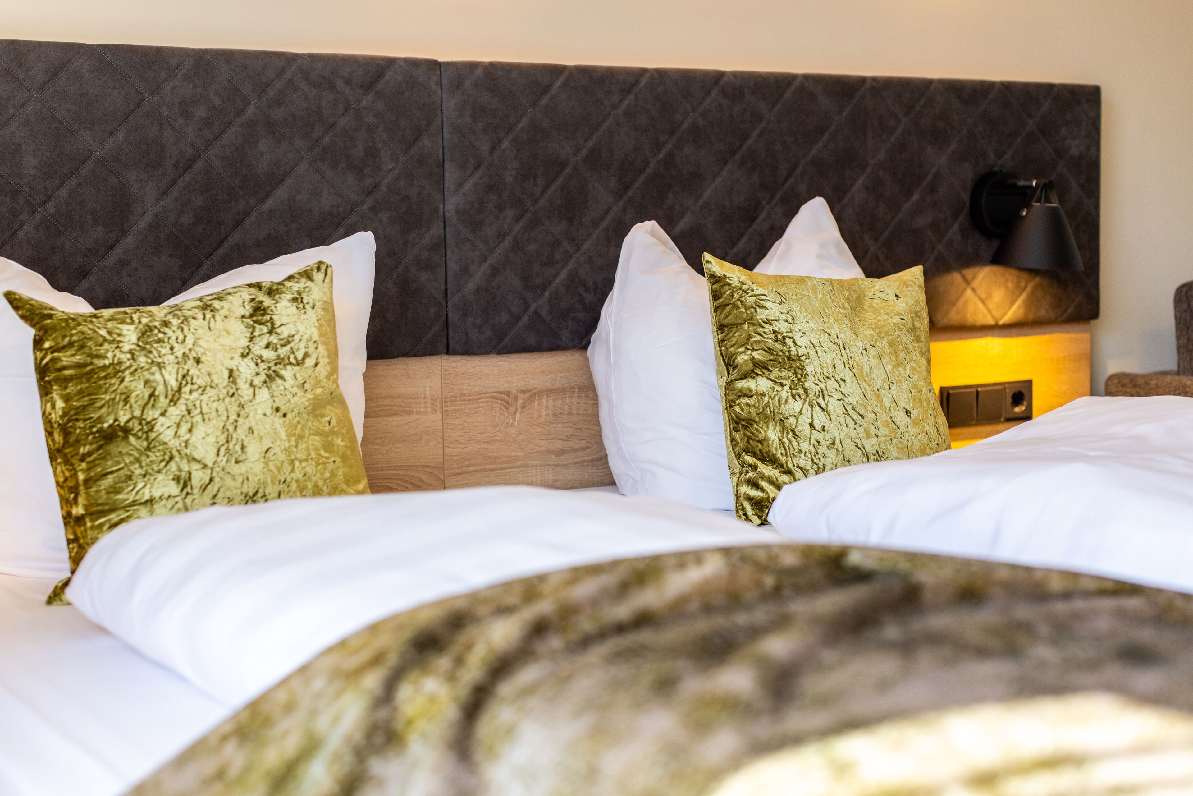A hotel room with a tufted headboard, a wooden bedside table, a lamp, and two beds with white and gold patterned pillows.