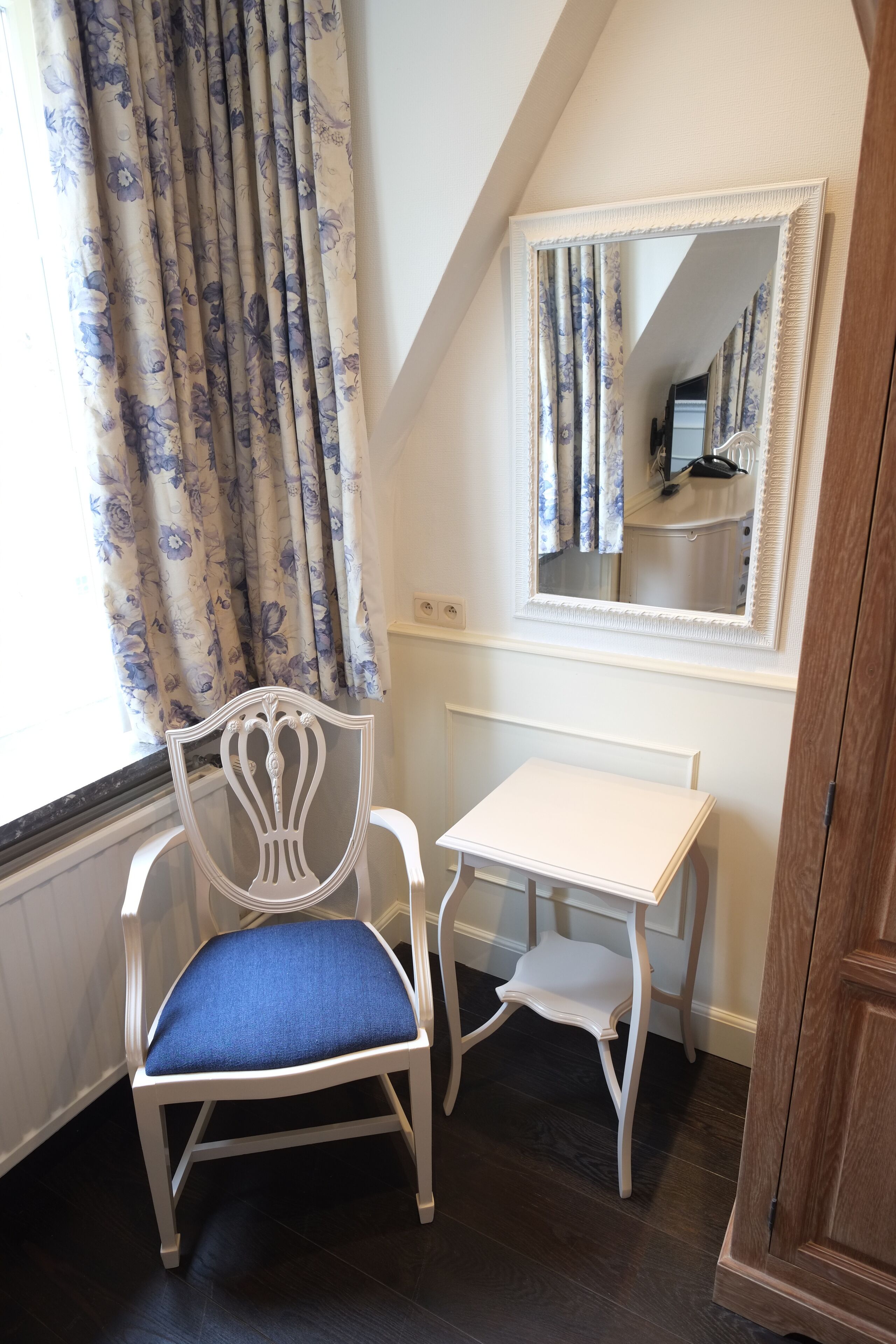A small room with a white chair with a blue cushion, a white table, a mirror on the wall, and a window with floral curtains.