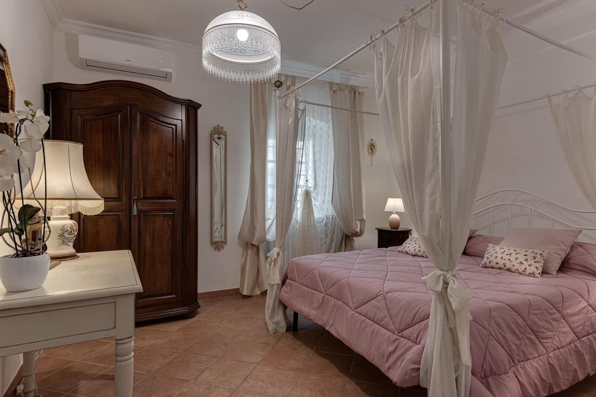 A bedroom with a four-poster bed, a wooden wardrobe, a bedside table with a lamp, and a chandelier.