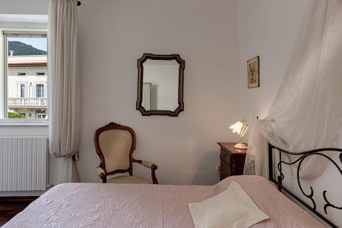 A bedroom with a bed, a chair, a nightstand, a mirror, and a window with curtains.