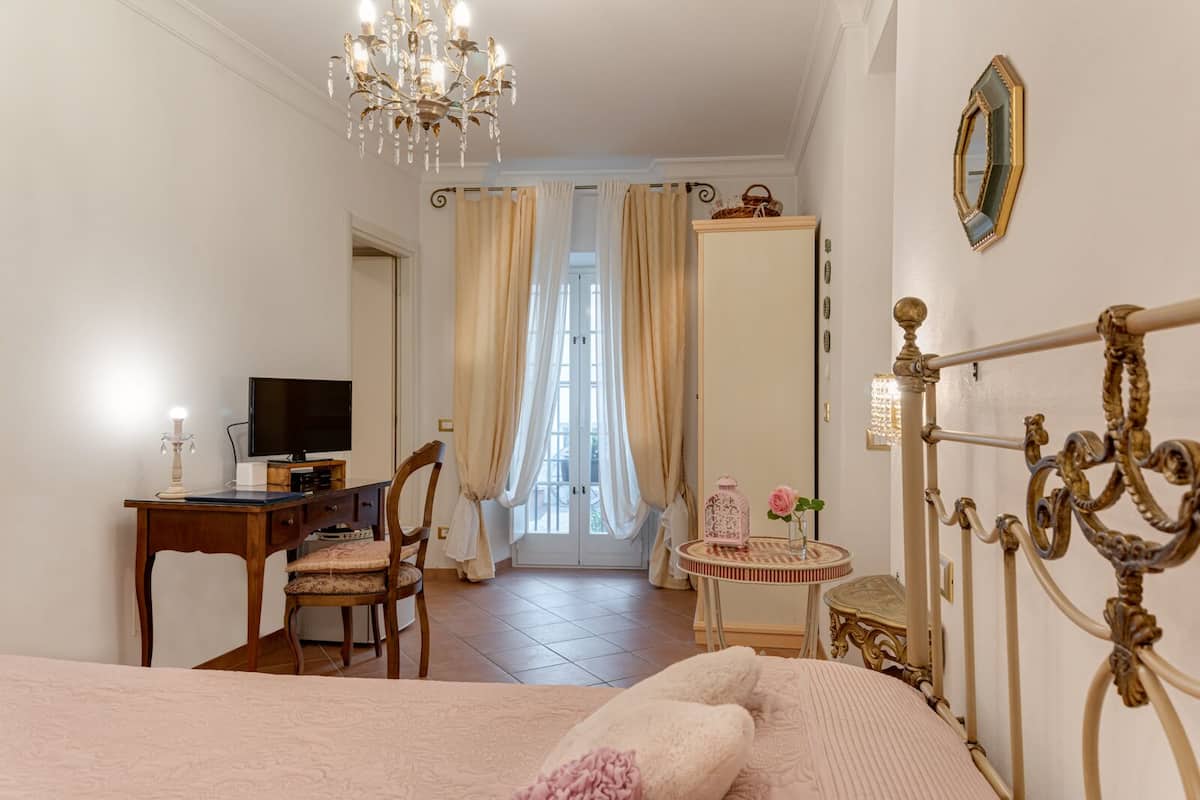 A bedroom with a bed, a desk with a TV, a chair, a chandelier, a mirror, and a door leading to a balcony.