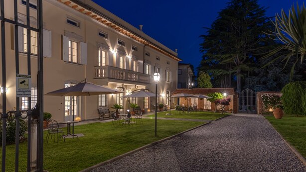 A well-lit outdoor area with a gravel path leading to a building with multiple windows and a balcony. There are outdoor tables and chairs under umbrellas, and a garden with various plants and a large tree. - Villa Del Papa (Lucca)