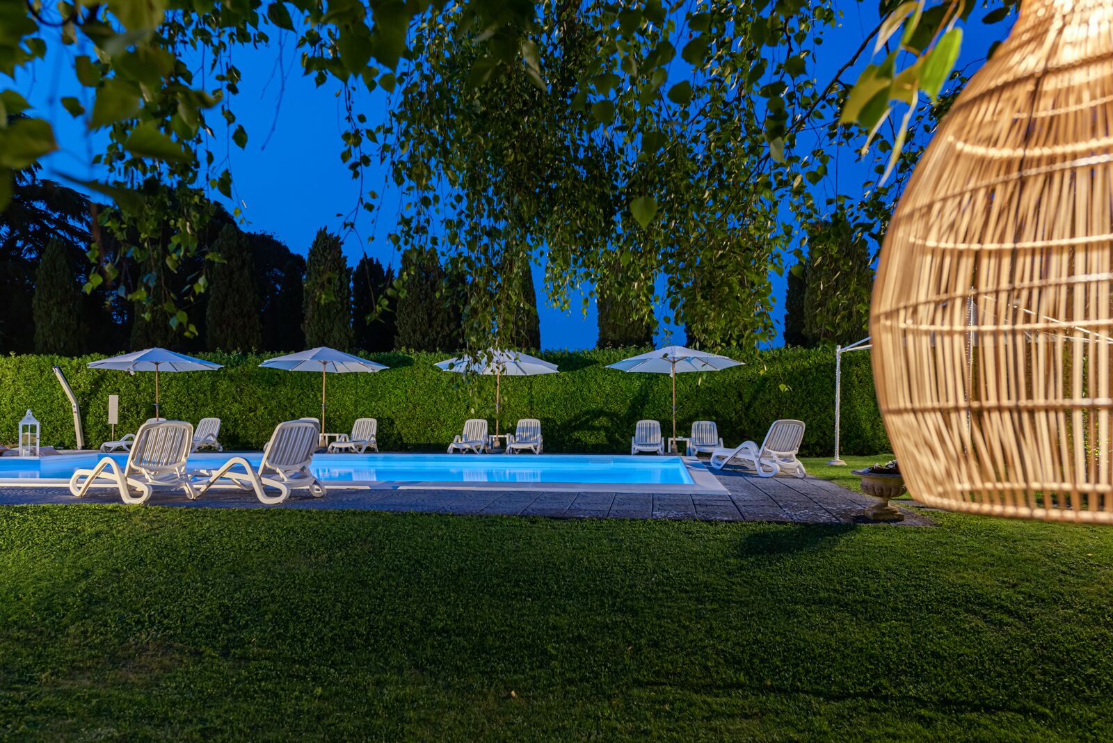 A poolside area with lounge chairs and umbrellas, surrounded by well-maintained hedges and trees.