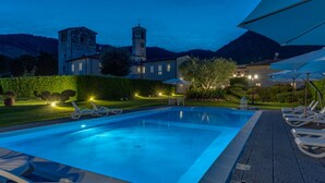 A swimming pool with lounge chairs and umbrellas, surrounded by well-maintained gardens and a historic building with a clock tower. - Villa Del Papa (Lucca)