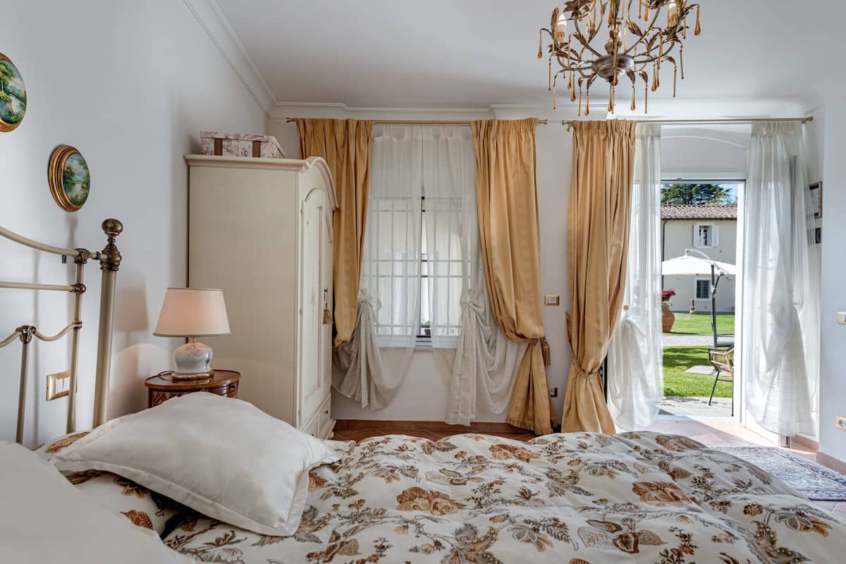 A bedroom with a bed, a nightstand, a lamp, a wardrobe, and a chandelier.