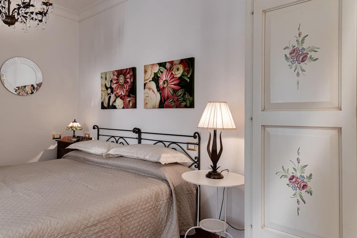 A bedroom with a bed, two wall-mounted paintings, a round mirror, a bedside table with a lamp, and a small round table with a lamp.