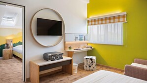 A hotel room with a bed, a desk with a suitcase, a mirror, a television, a window with curtains, and a wall with a checkered pattern. - Hey Lou Hotel Piding (Piding)