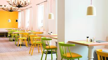 A brightly lit dining area with yellow and green chairs, white tables, and a chandelier.