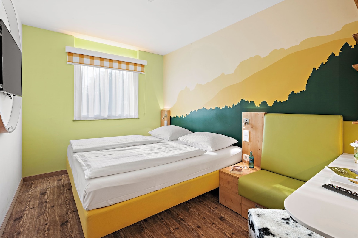 A room with a bed, a desk, and a chair. There is a window with curtains, a wall clock, and a wall-mounted TV.
