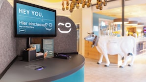 A reception desk with a digital screen displaying a greeting in German, a charging station, and a small animal statue in the background. - Hey Lou Hotel Piding (Piding)