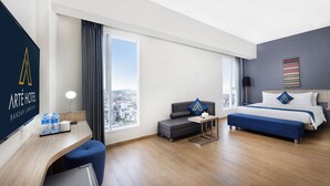 A hotel room with a desk, a bed, a sofa, and a large window offering a city view.