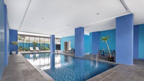 An indoor swimming pool with blue walls and a tiled floor, surrounded by lounge chairs and potted plants.