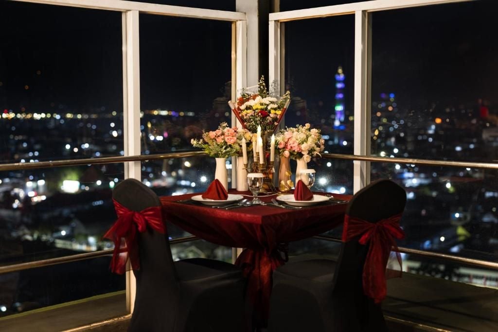 Mesa para dos con mantel rojo, velas y flores, con vista a un paisaje urbano nocturno.