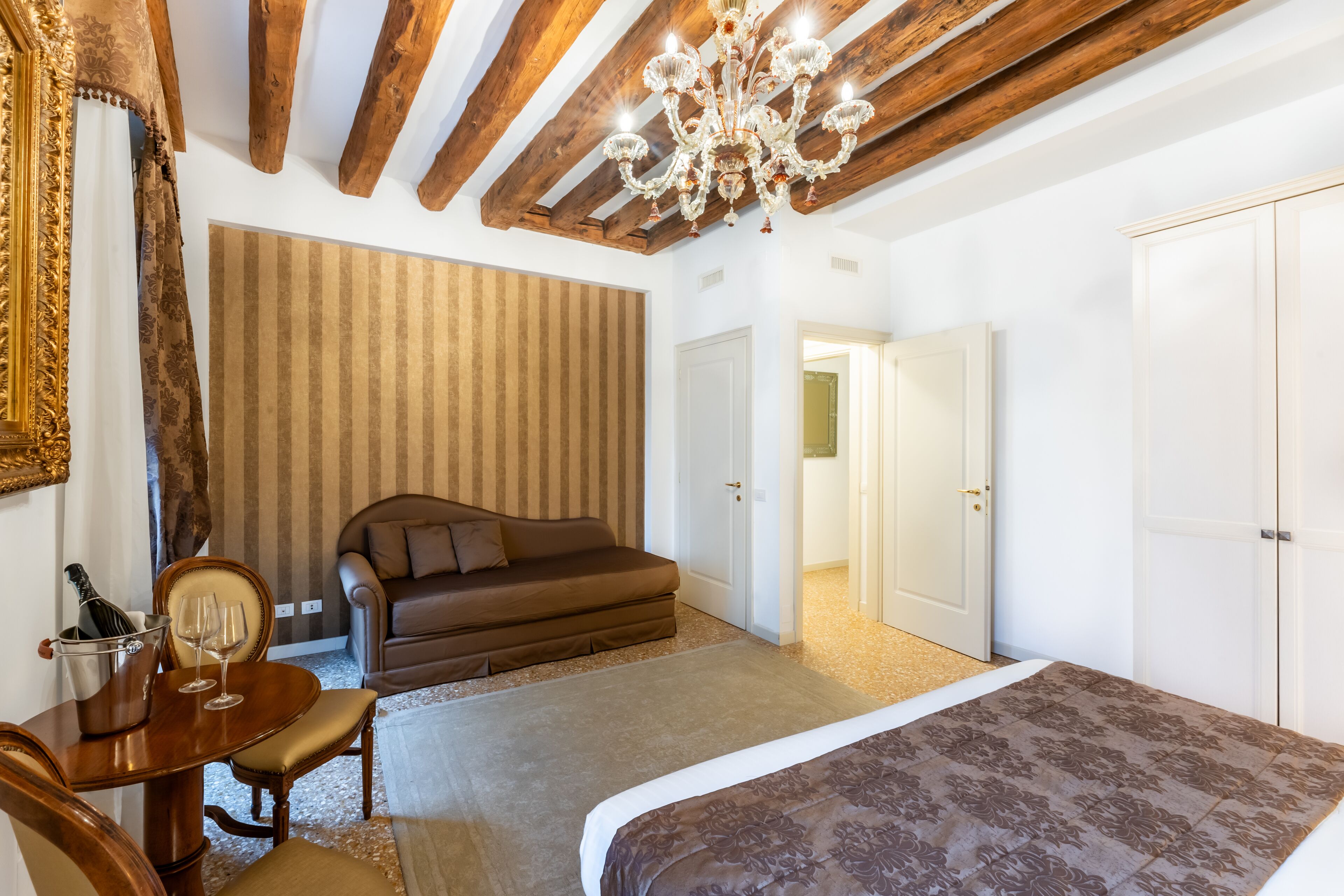A bedroom with a bed, a wooden table with a bottle and glasses, a brown sofa, and a chandelier.