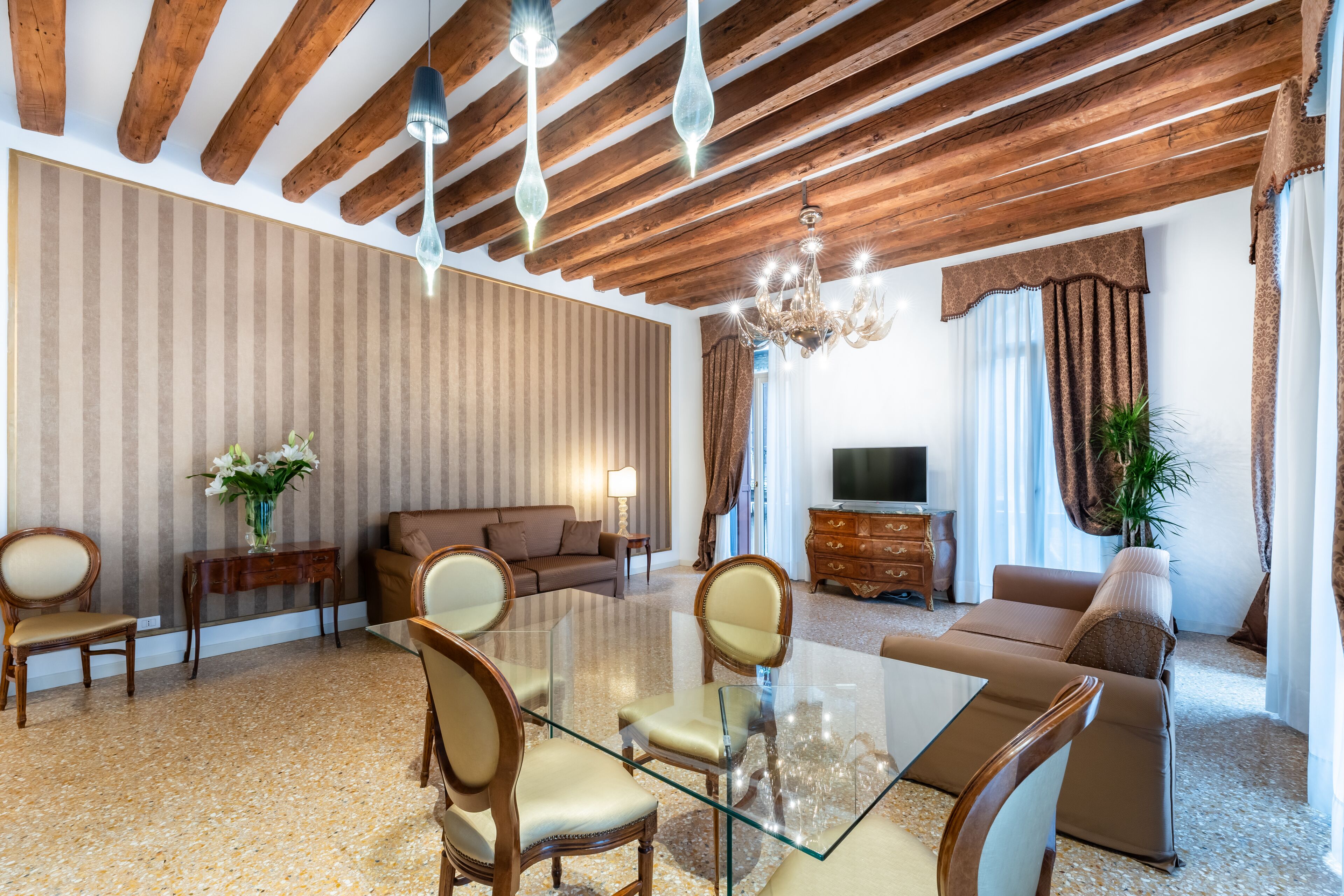 A modern living room with a glass dining table, wooden ceiling beams, and a sofa set.