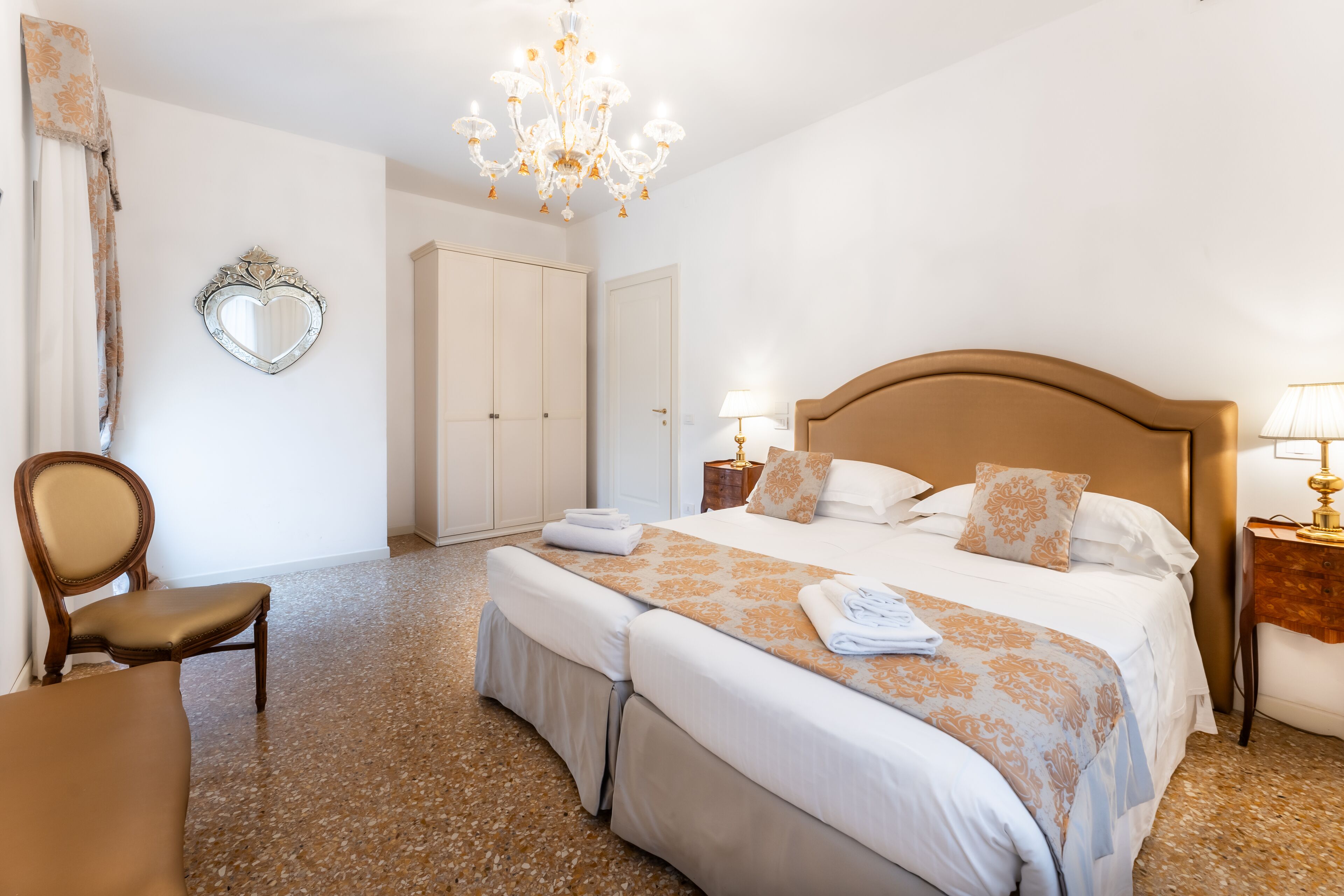 A hotel room with a large bed, two bedside tables, a chandelier, a mirror, and a wardrobe.