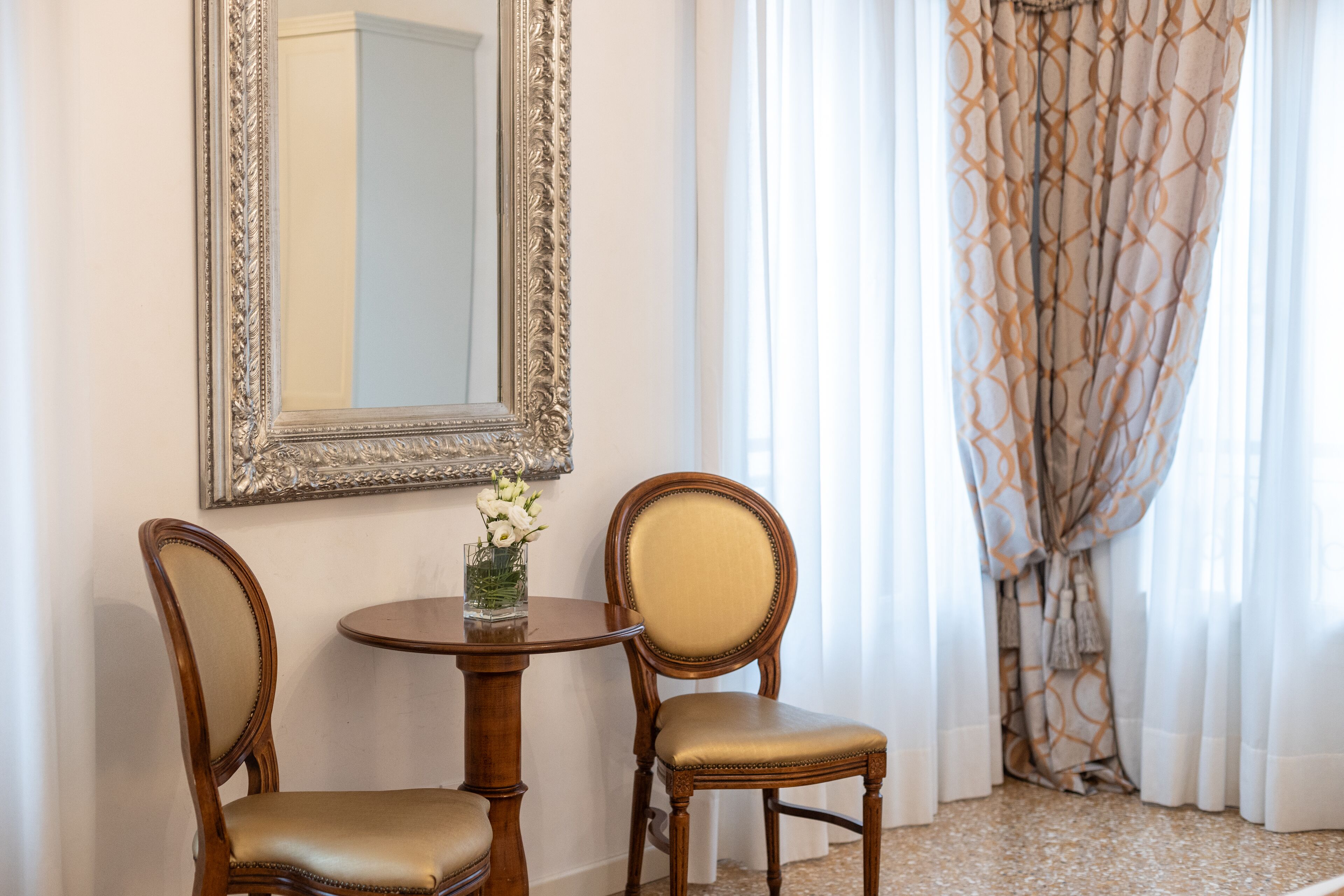 A room with two wooden chairs, a small round table with a vase of flowers, and a large mirror on the wall.