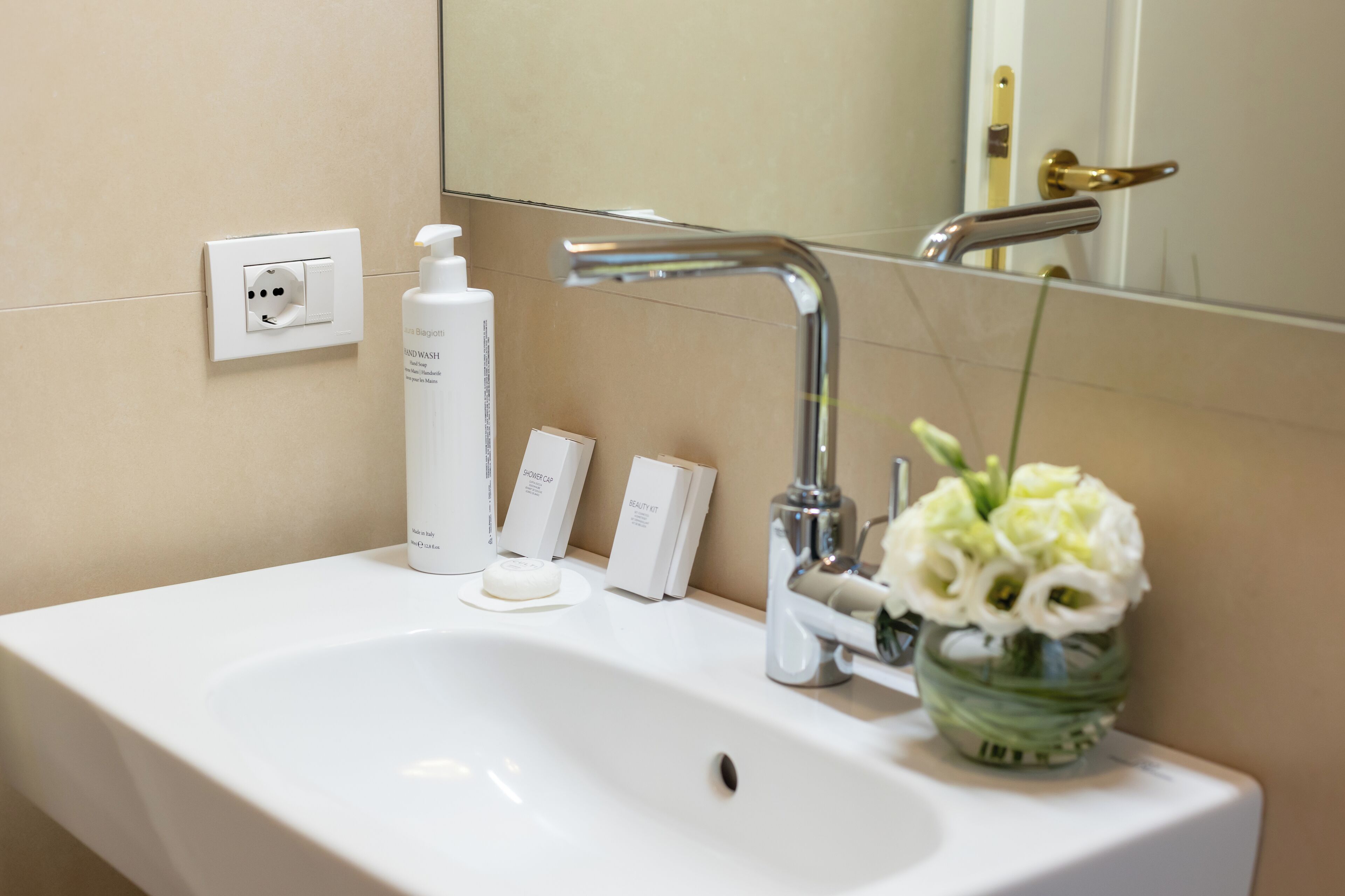 Un baño con un lavabo blanco, un espejo, un grifo, un florero con flores y artículos de aseo en la encimera.