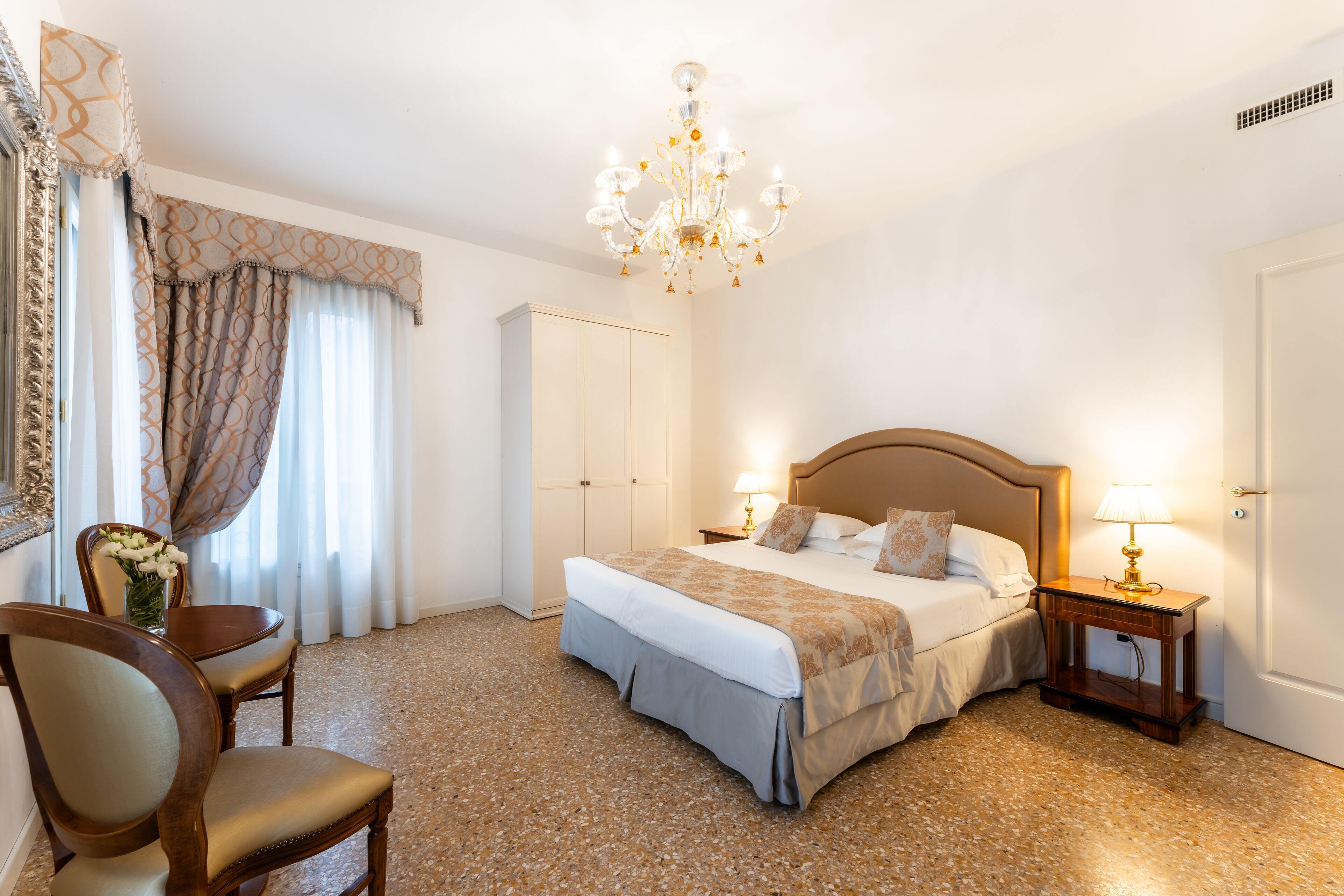 Una habitación de hotel con una cama grande, mesitas de noche con lámparas, una silla, un escritorio y una araña.