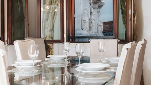 Una mesa de comedor dispuesta con platos blancos, vasos y cubiertos, rodeada de sillas color beige, con vista a una escultura a través de una ventana.