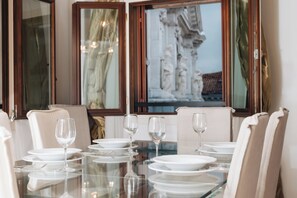 Een eettafel gedekt met witte borden, glazen en bestek, omringd door beige stoelen, met uitzicht op een sculptuur door een raam.