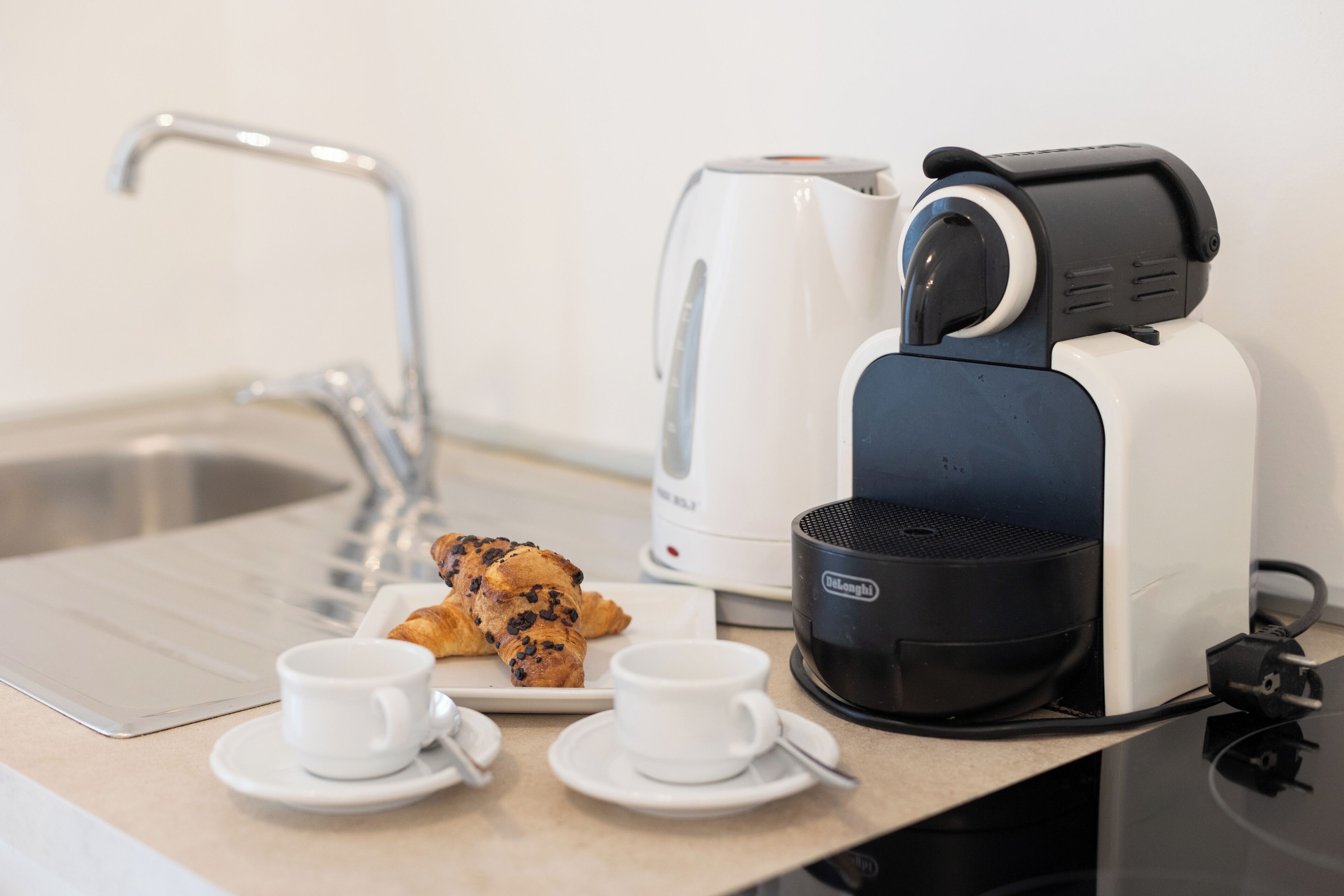 A kitchenette with a coffee maker, a kettle, and croissants on a countertop.