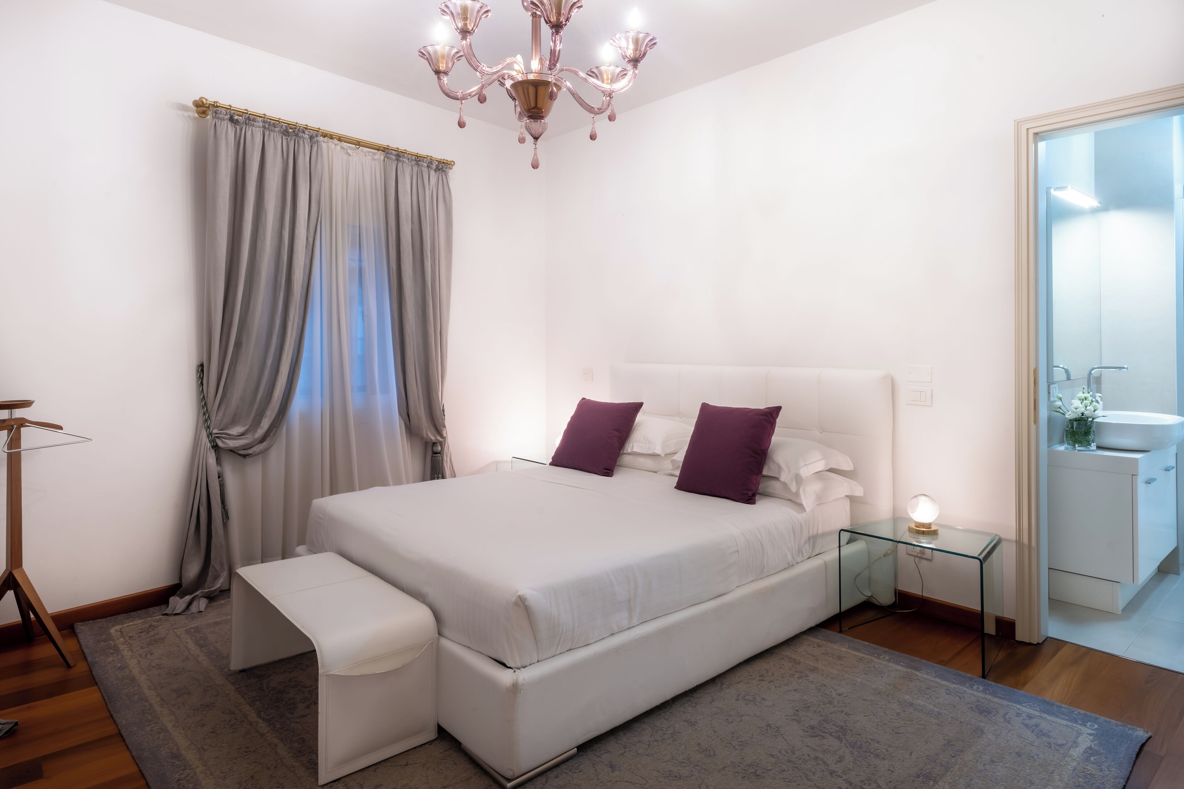 A modern bedroom with a large bed, bedside tables, a bench, a chandelier, and a bathroom visible through an open door.