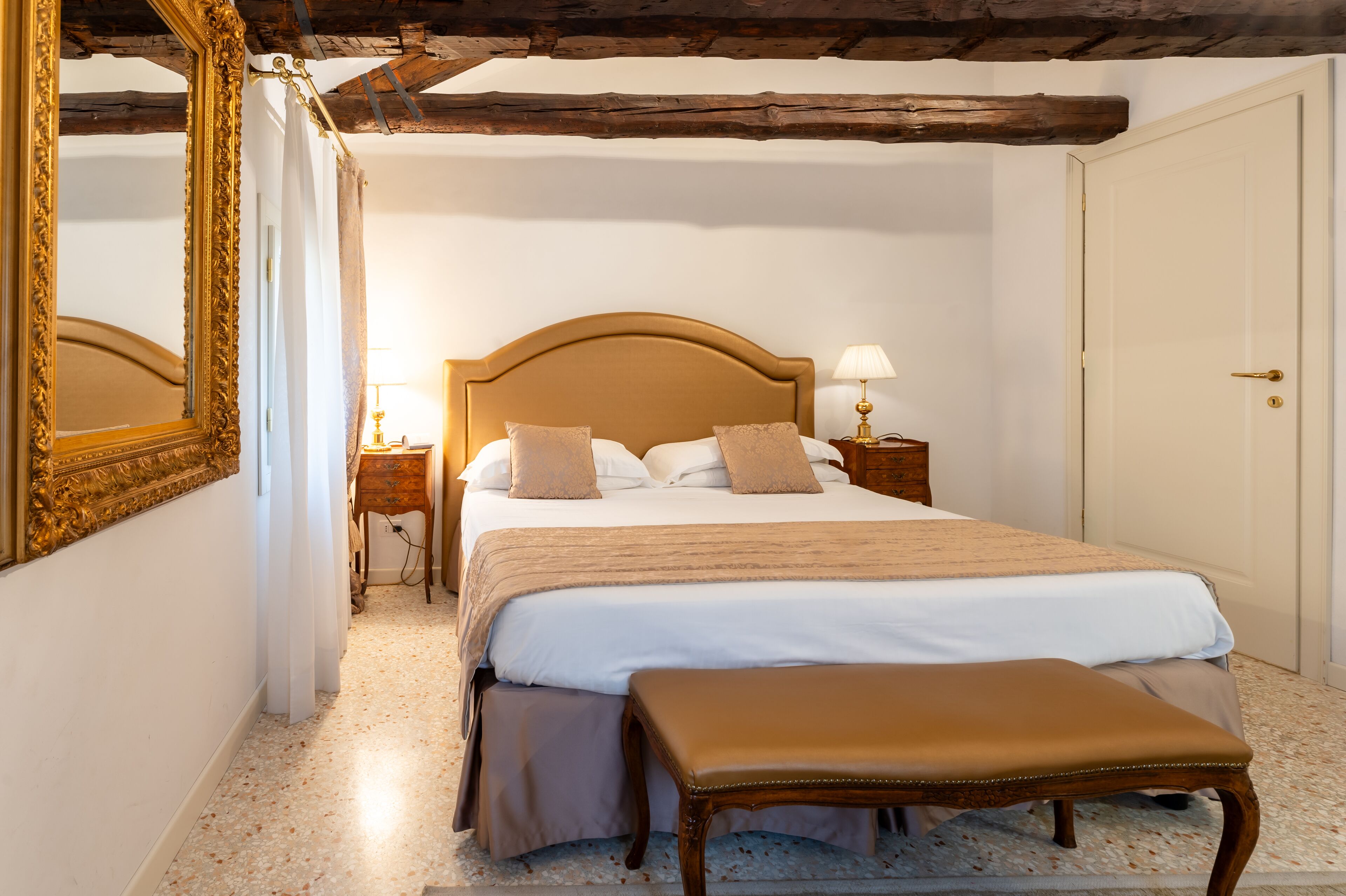 Quarto com uma cama grande, um banco de madeira, uma mesinha de cabeceira com abajur e uma porta.