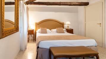 Quarto com uma cama grande, um banco de madeira, uma mesinha de cabeceira com abajur e uma porta.