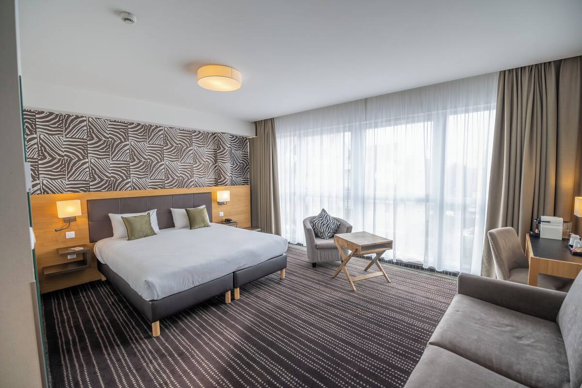 Ein Hotelzimmer mit einem großen Bett, einem kleinen Tisch, einer Couch und einem Sessel. Es gibt ein Fenster mit Vorhängen und einer Wand mit Muster.