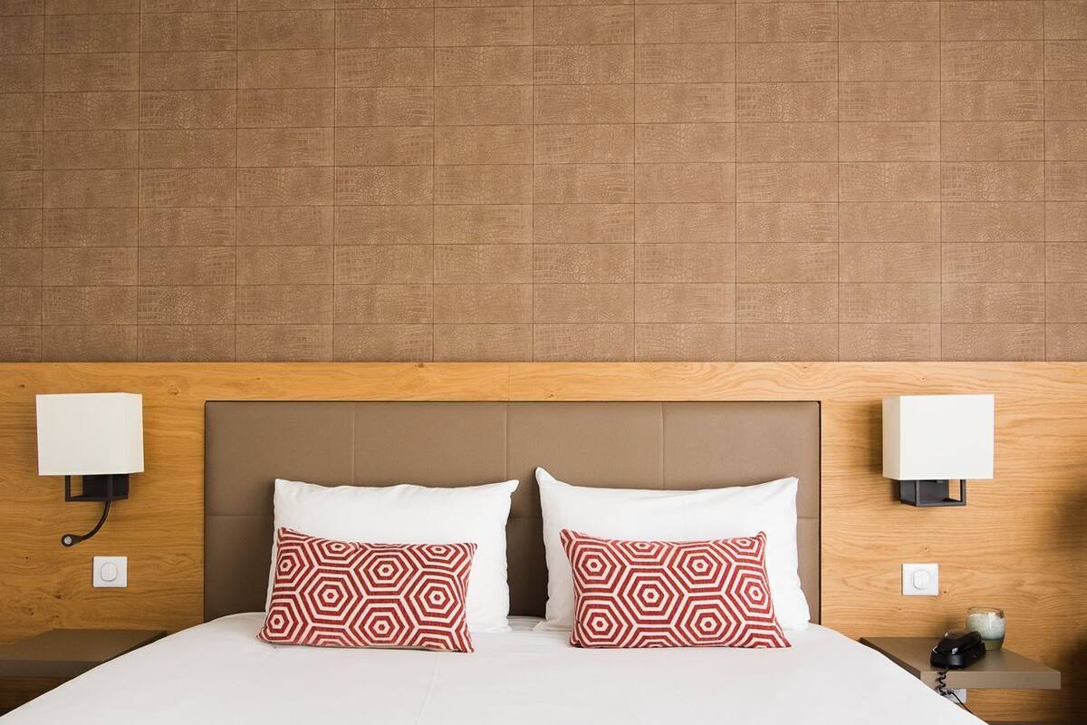 Ein Hotelzimmer mit hölzernem Kopfteil, zwei Nachttischlampen, einem Bett mit weißer Bettwäsche und zwei gemusterten Kissen.