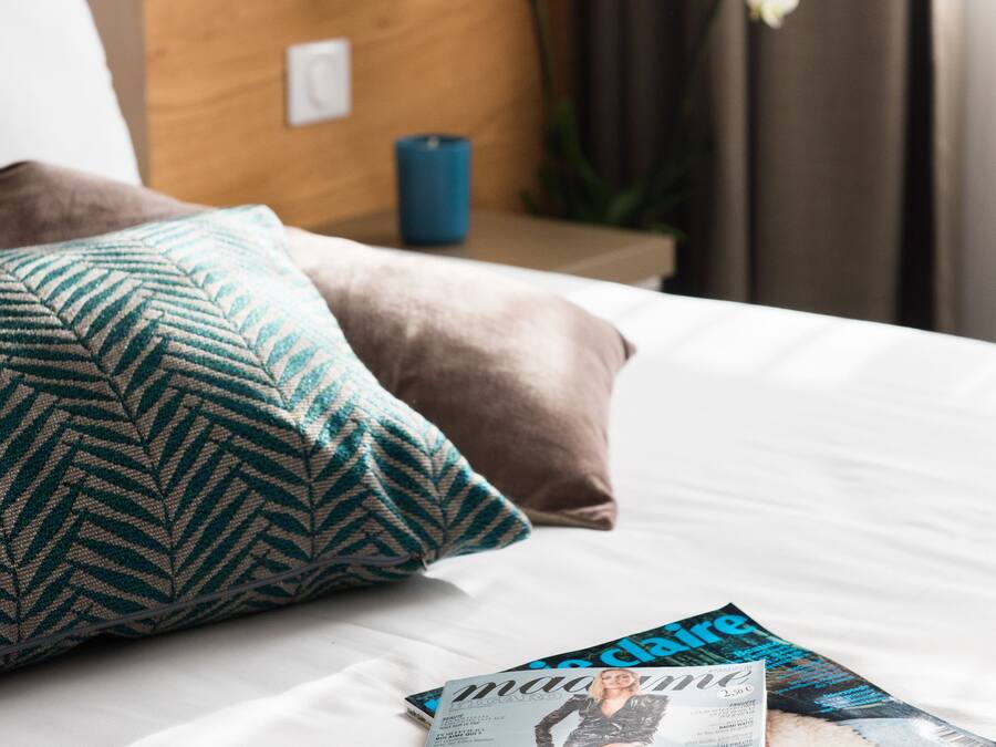 A neatly made bed with patterned pillows, a magazine, and a wall-mounted lamp.