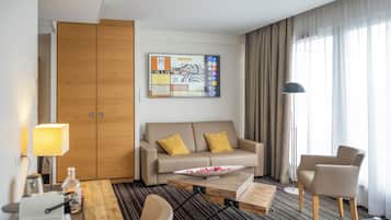 Habitación de hotel con sofá, una mesa de centro, escritorio con lámpara, silla y ventana grande con cortinas.