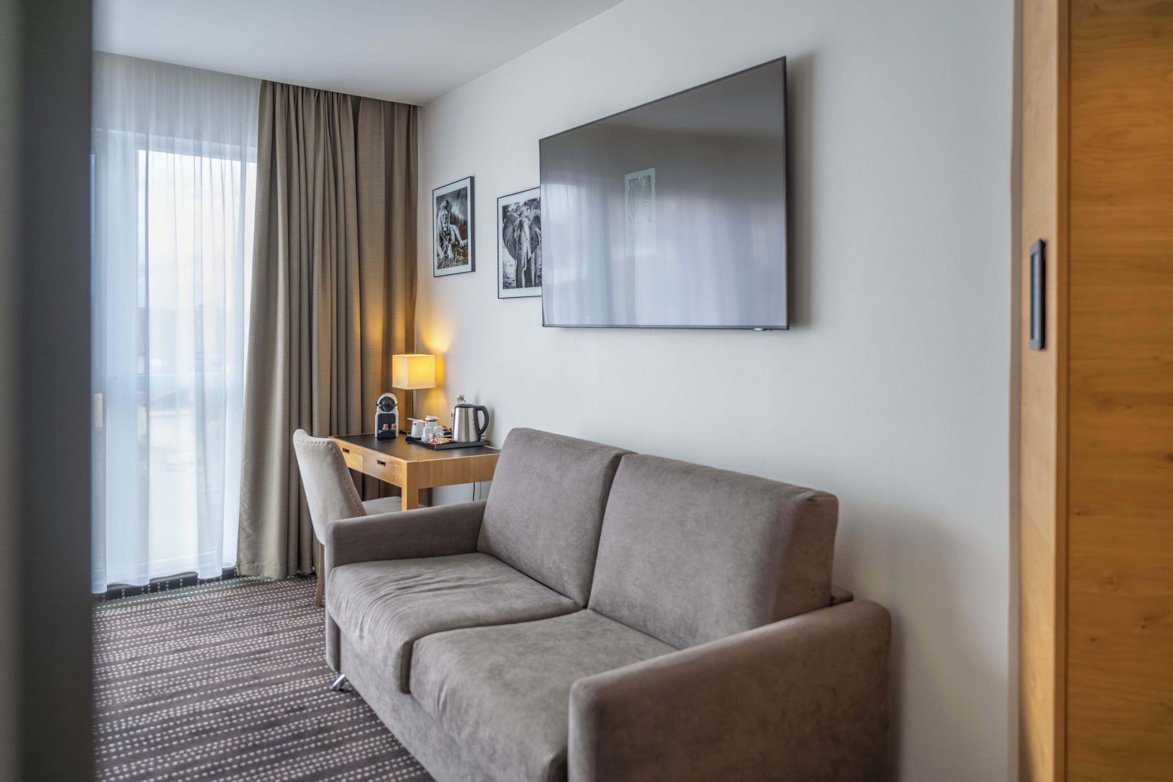 A hotel room with a sofa, a desk with a lamp, and a flat-screen TV mounted on the wall.