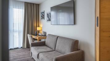 A hotel room with a sofa, a desk with a lamp, and a flat-screen TV mounted on the wall.