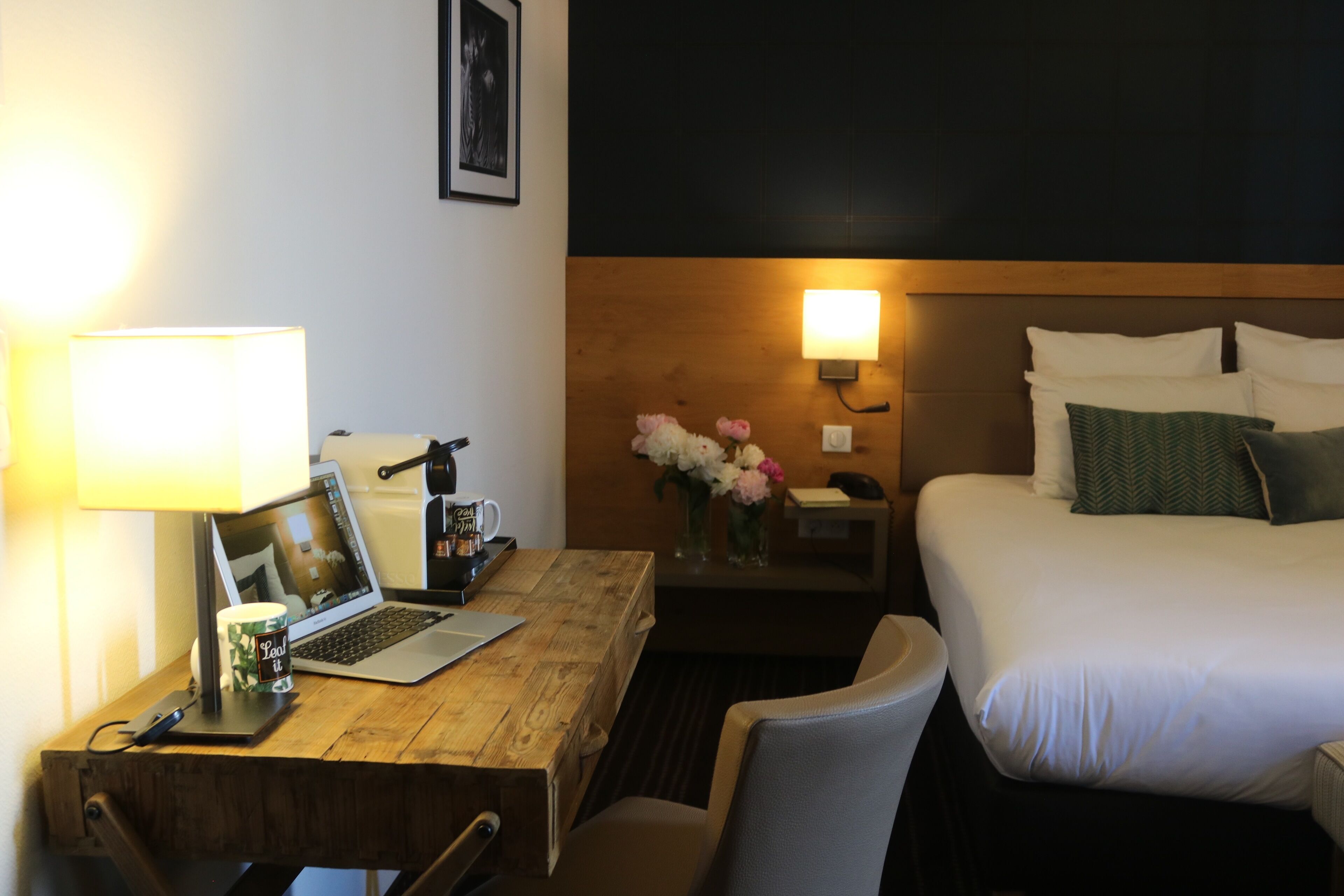 Habitación de hotel con un escritorio de madera, una computadora portátil, una lámpara, una mesita de noche con un jarrón de flores, una cama con almohadas y una lámpara de pared.