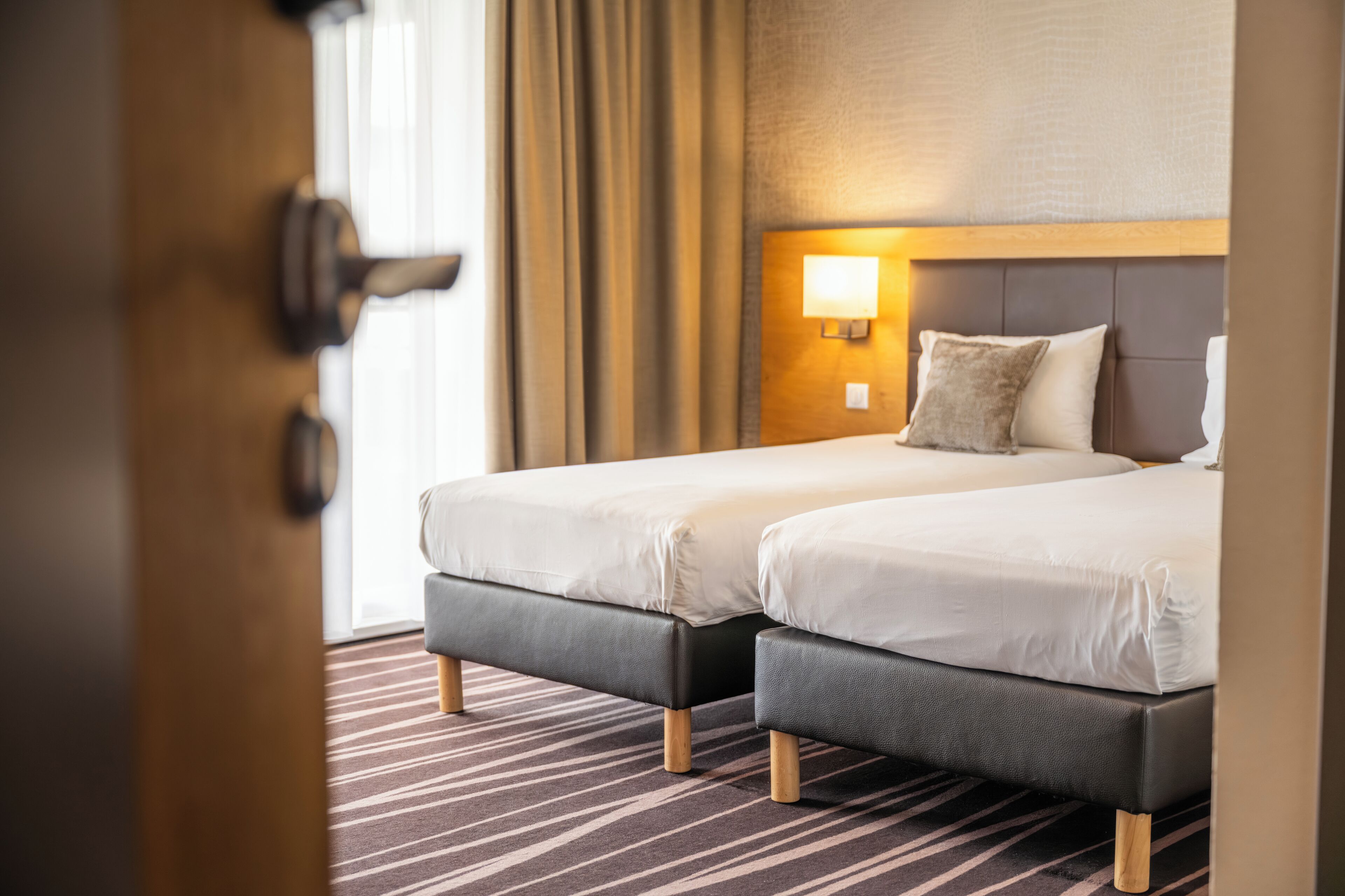 A hotel room with two beds, a wooden headboard, a nightstand with a lamp, and a striped carpet.