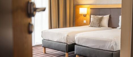 A hotel room with two beds, a wooden headboard, a nightstand with a lamp, and a striped carpet.