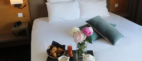 A neatly made hotel bed with white linens, a tray with croissants, a cup of coffee, a vase with flowers, and a teapot.