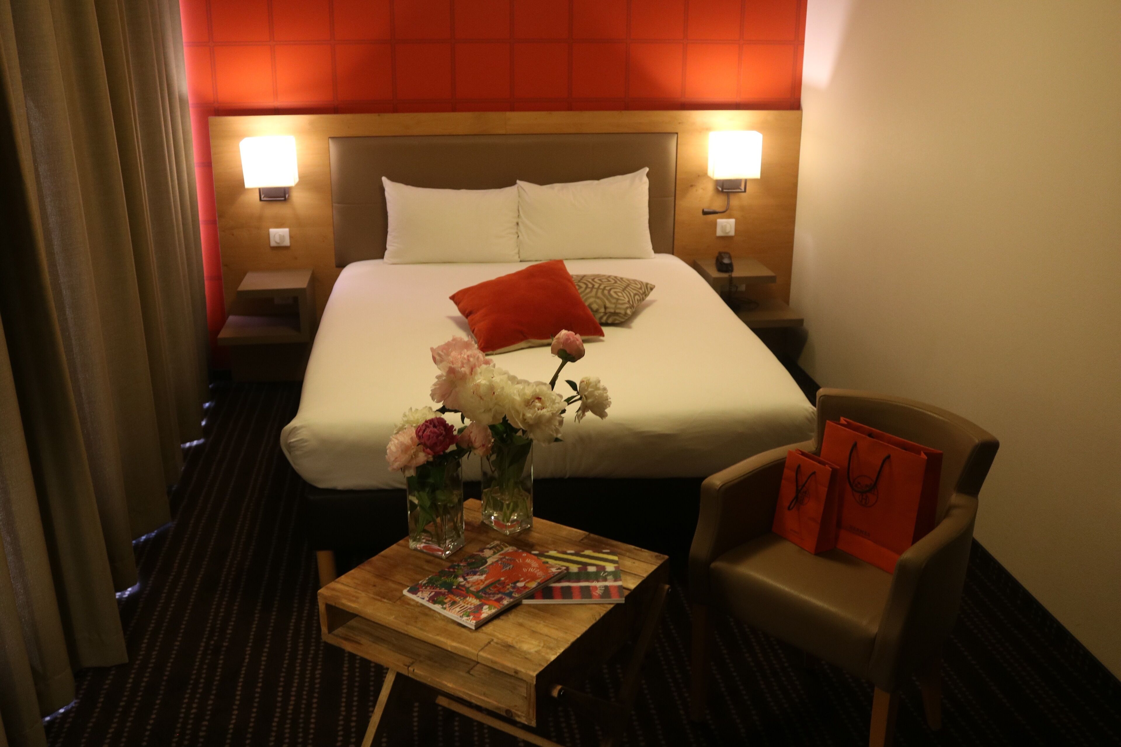 Une chambre d’hôtel avec un lit, des lampes de chevet, une table de chevet, une table en bois avec un magazine et un vase de fleurs, et une chaise sur laquelle sont posés des sacs de shopping.