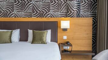 A hotel room with a large bed, two pillows, a nightstand with a lamp, and a telephone. The room has a zebra-patterned wallpaper and a wooden headboard.