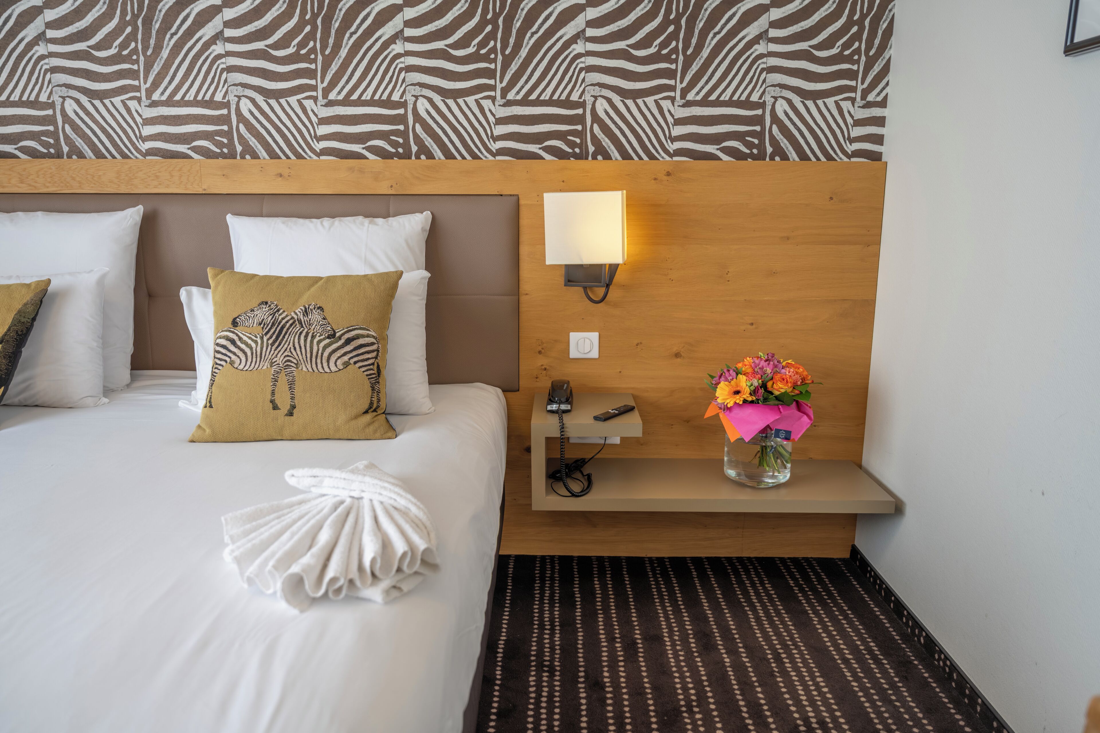 A hotel room with a zebra-patterned headboard, a bedside table with a lamp, a phone, and a vase of flowers.