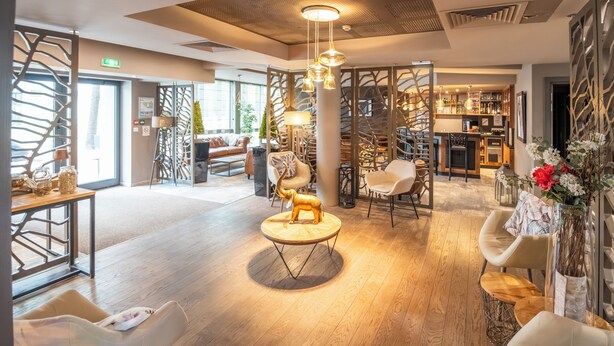 A modern hotel lobby with a wooden floor, a round coffee table with a golden elephant sculpture, and a bar area with a staircase. - Brit Hotel Le Lodge (Strasbourg)
