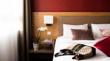 Une chambre d’hôtel bien rangée, avec une tête de lit en bois, du linge de lit blanc, un coussin rouge, une lampe blanche et une petite table sur laquelle se trouvent un magazine et une orchidée en pot.