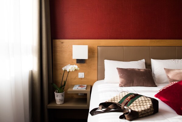 A neatly made hotel room with a wooden headboard, white bedding, a red pillow, a white lamp, and a small table with a magazine and a potted orchid.