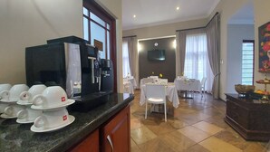 Quarto de hotel com cafeteira, xícaras de chá, área de refeições com mesas e cadeiras e uma TV na parede.
