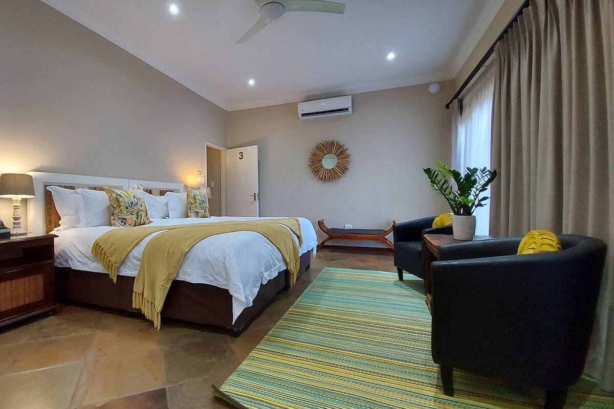 Ein Hotelzimmer mit einem großen Bett, einem Schreibtisch mit Lampe, einem Sessel, einer Zimmerpflanze und Blick ins Freie.