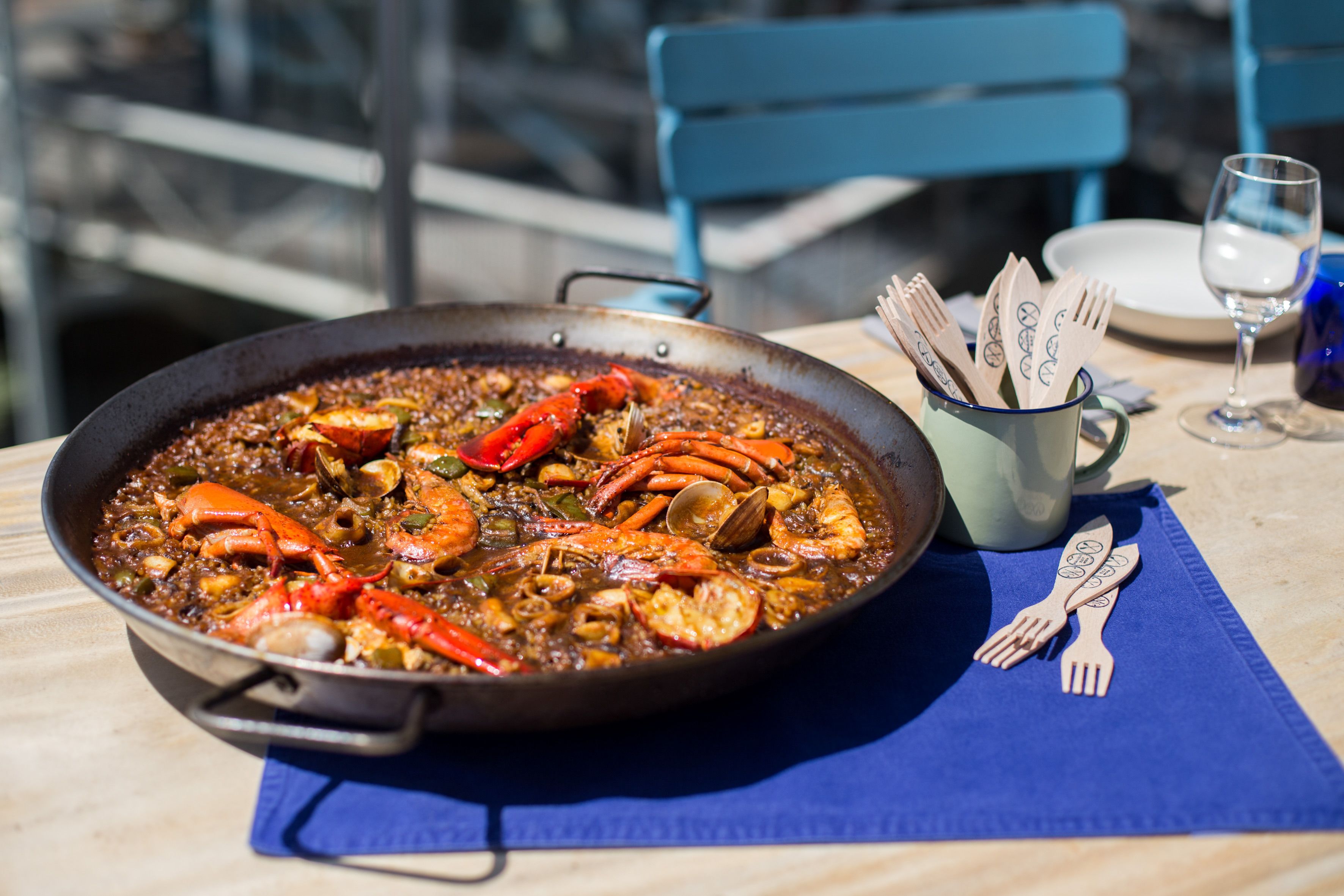 Una paella de mariscos en una sartén sobre una mesa con un mantel azul, cubiertos de madera y un vaso de agua.